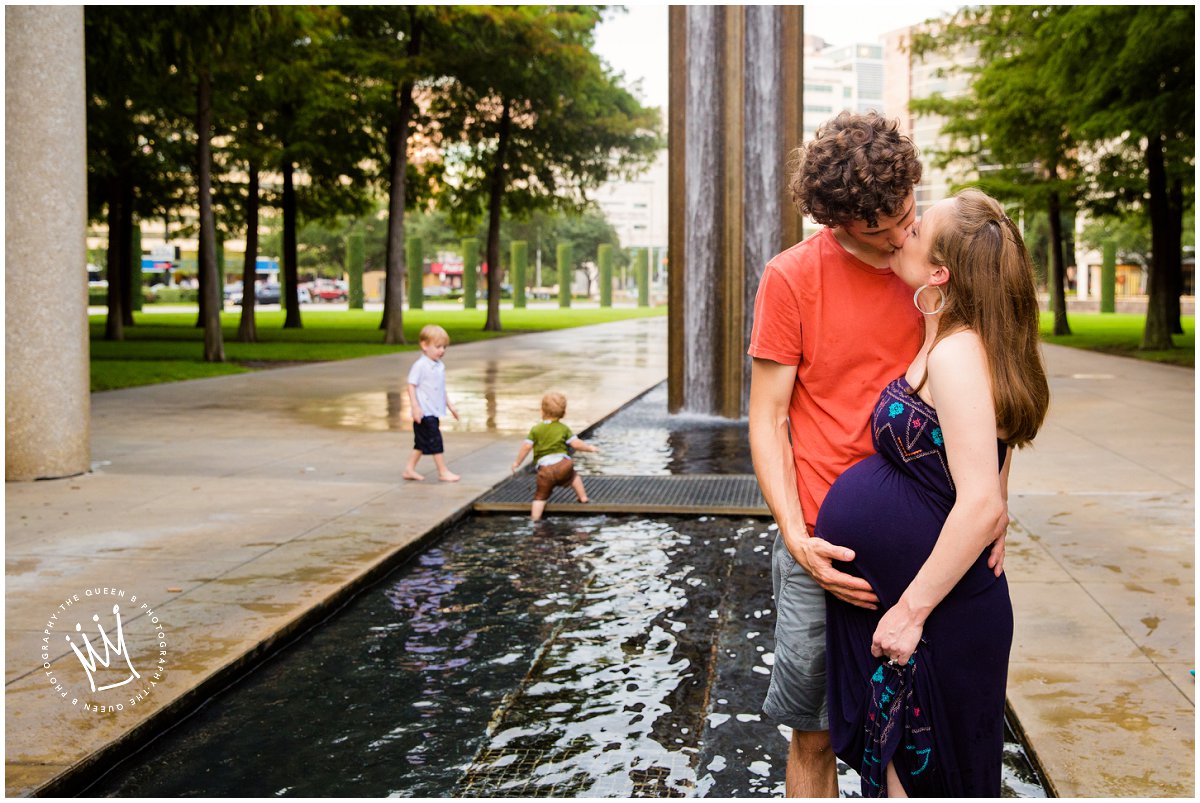 kissing expectant couple little boys play in water fountain captured by Houston Maternity Photographer
