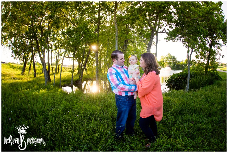 family of three with baby girl in the grass by the bank of the lake at sunset
